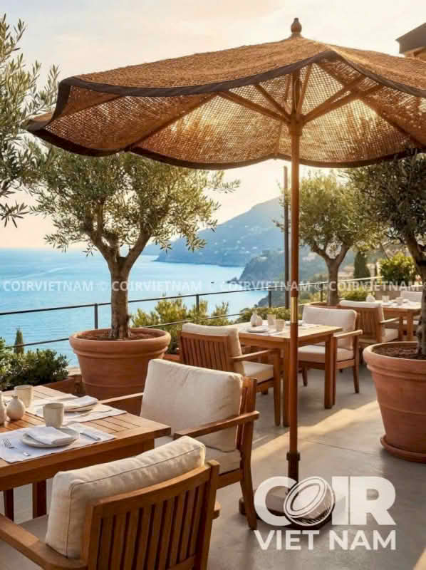 Outdoor patio seating area with wooden tables and chairs, large woven beach umbrellas, potted olive trees, and a scenic ocean view in the background.