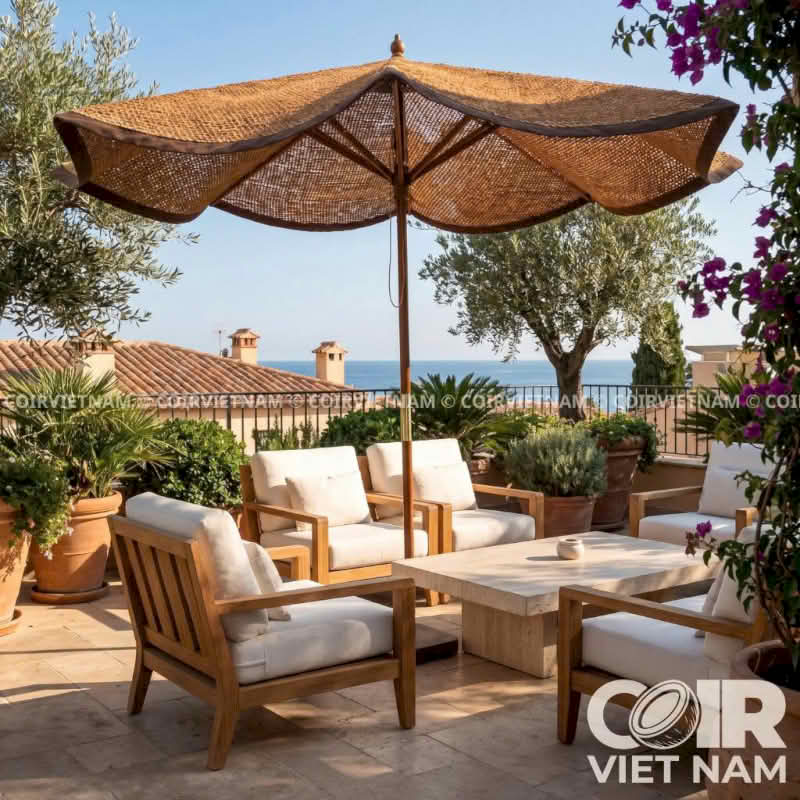 Outdoor patio furniture set with a large woven wicker umbrella, wooden armchairs with off-white cushions, and a matching stone coffee table, set against a scenic background of the sea and sky.