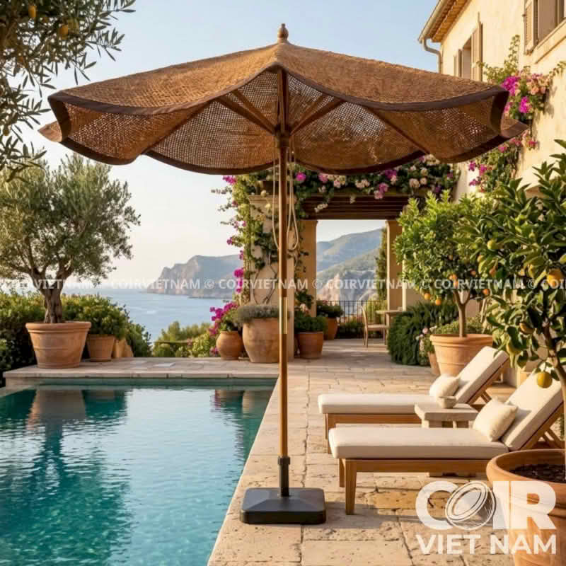 A large woven wicker patio umbrella with a square black weighted base stands beside a swimming pool on a stone terrace. The scene includes two cream lounge chairs, potted plants, a building exterior, and distant sea views.