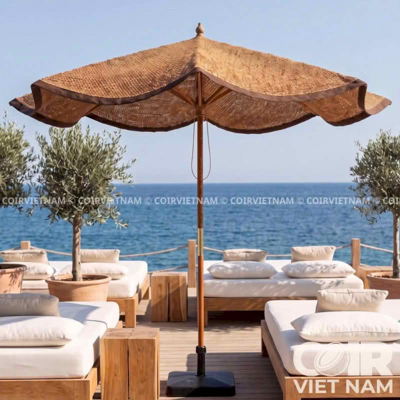A large woven straw beach umbrella with a black base stands on a wooden deck overlooking the blue sea. Comfortable white cushioned lounge chairs and potted trees are arranged around it in an outdoor seaside setting.