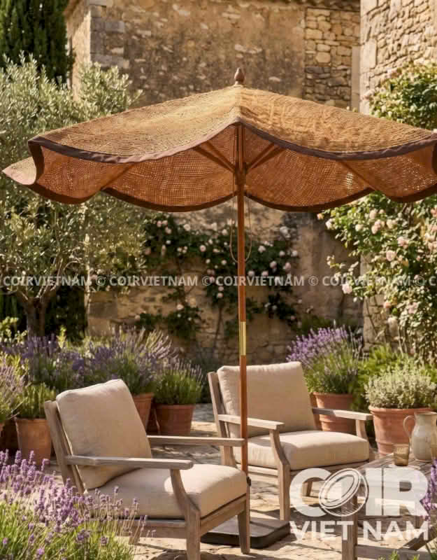 A large woven patio umbrella in a rustic garden setting, shading two wooden lounge chairs with beige cushions. The background features a stone wall and potted lavender plants.