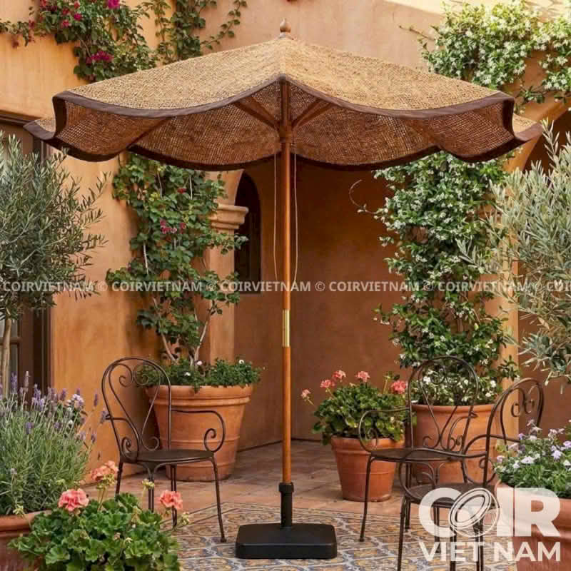 A large outdoor patio umbrella with a woven straw-colored canopy and dark brown trim, set in a Mediterranean-style courtyard with potted plants, metal chairs, and a terracotta wall. The umbrella has a wooden pole and a square black base.
