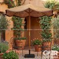 A large outdoor patio umbrella with a woven straw-colored canopy and dark brown trim, set in a Mediterranean-style courtyard with potted plants, metal chairs, and a terracotta wall. The umbrella has a wooden pole and a square black base.